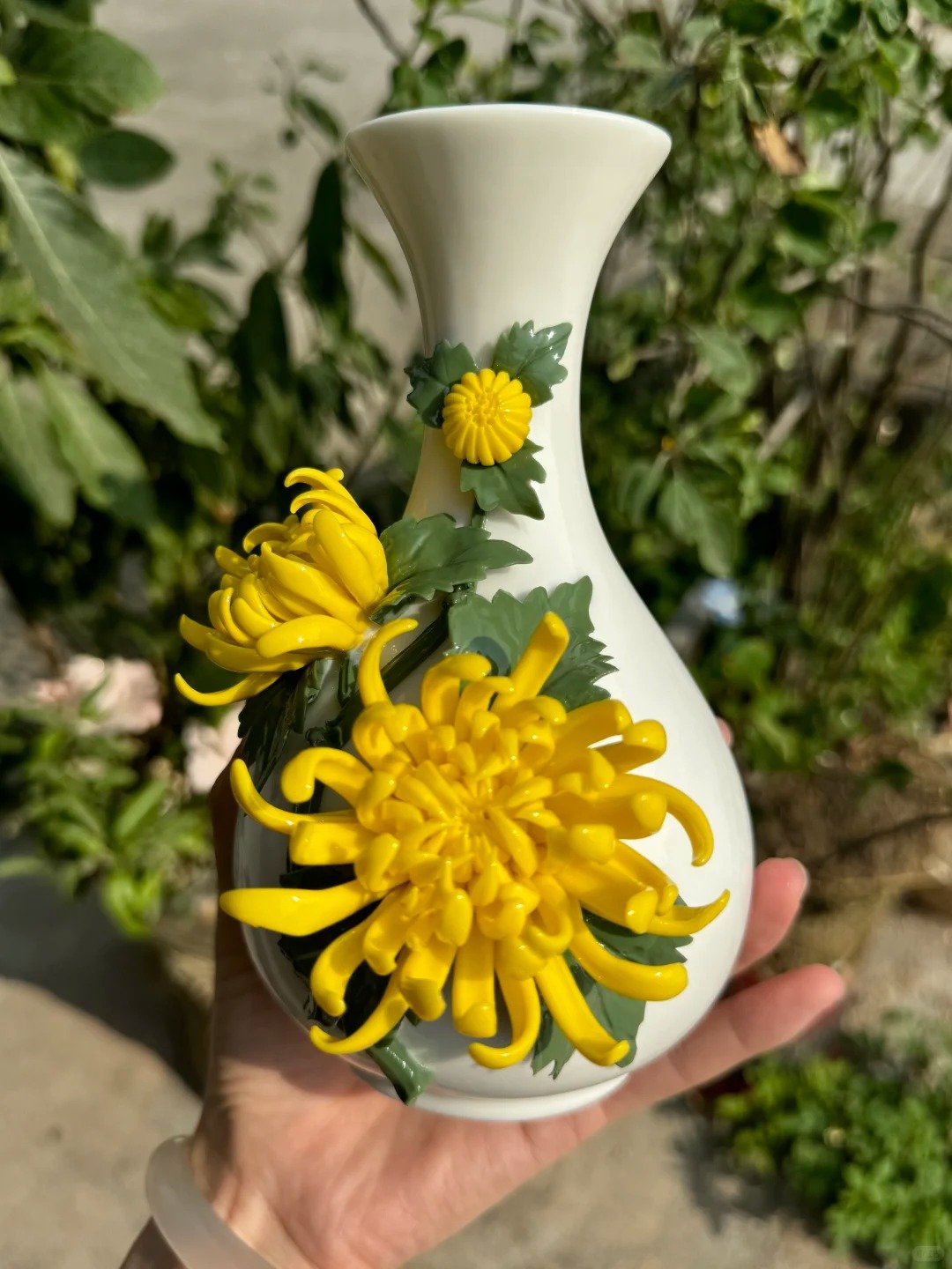 Hand holding a white porcelain vase decorated with yellow chrysanthemum ceramic flowers and green leaves