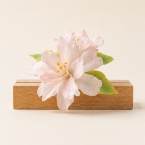 Pale pink begonia ceramic flower with green leaves on a wood base against a clean studio background